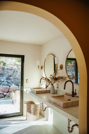 A bathroom with a large arched doorway, two sinks with gold fixtures, a mirror, and a window overlooking a rocky landscape. - OESTE Desert Escape in Morongo Valley - Central AC, 5 private Acres, Starlink (Morongo Valley)