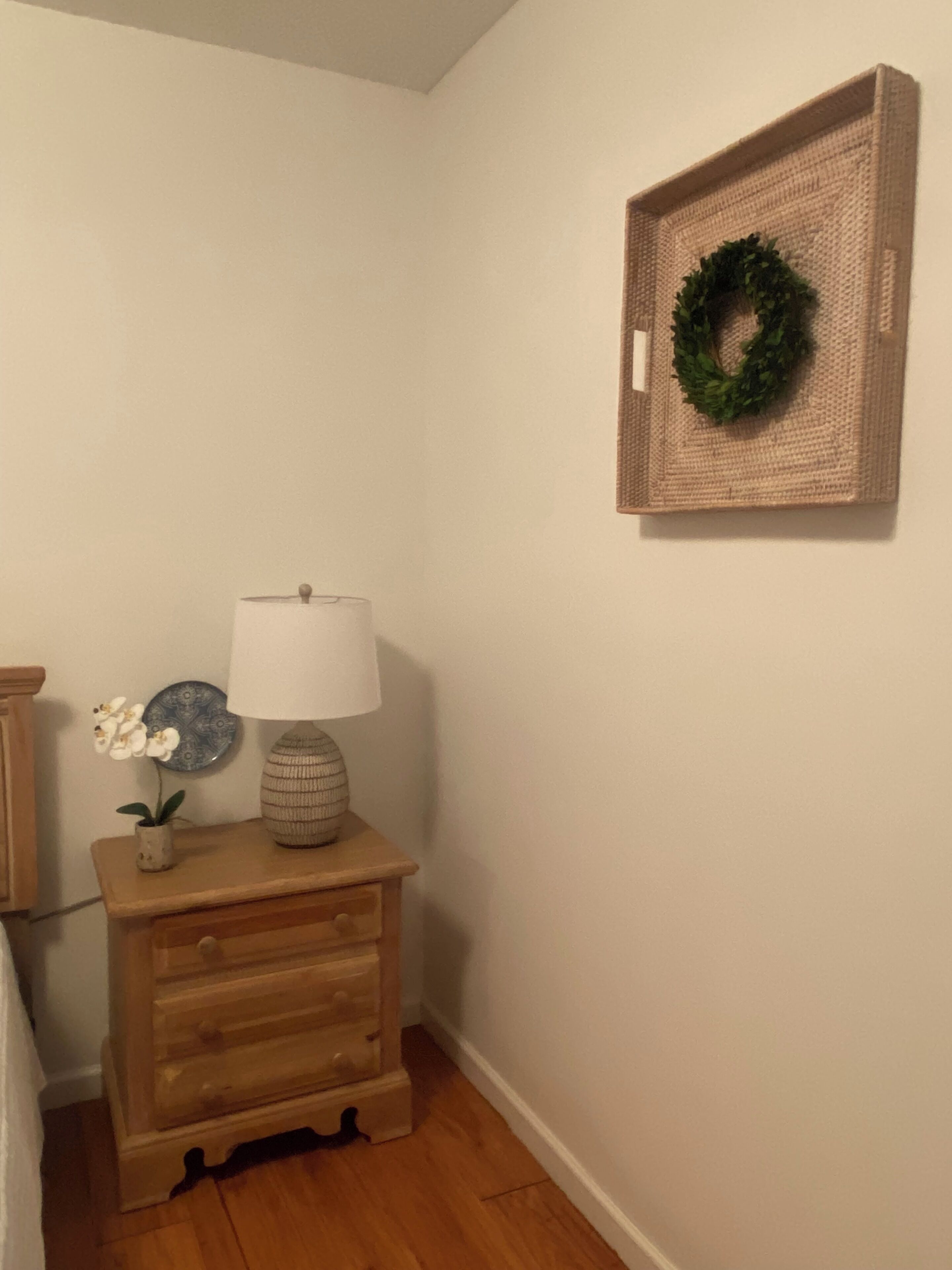 A corner with a wooden nightstand, a lamp, a small vase with flowers, a decorative plate, and a woven wall hanging.