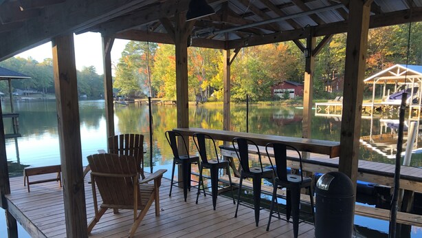 A wooden deck with a table and chairs overlooking a calm lake, surrounded by trees and a dock. - Waterfront cabin on Lake Martin near restaurants, golf and, AU football (Dadeville)