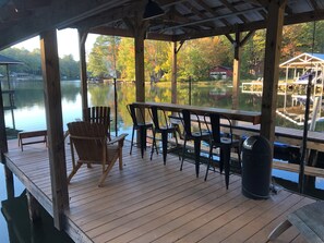 A wooden deck with a table and chairs overlooking a calm lake, surrounded by trees and a dock.