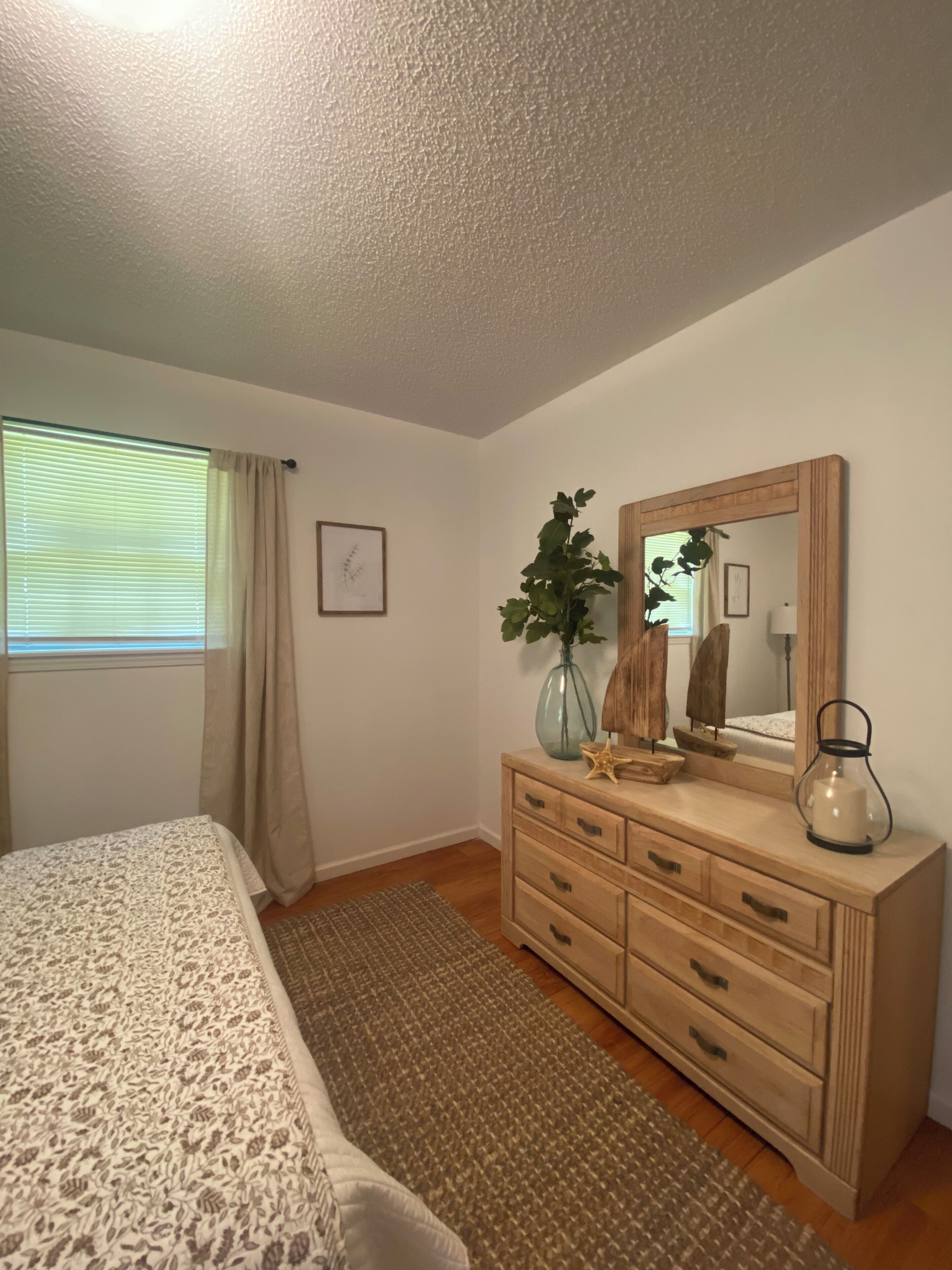 A bedroom with a bed, dresser, mirror, window with curtains, and a plant.