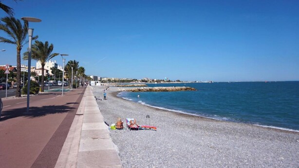 Une plage de galets avec une promenade dallée, des palmiers et un ciel bleu azur.