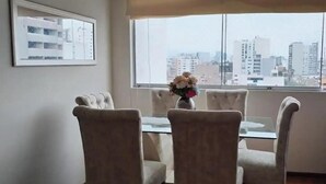 A dining area with a glass table and four upholstered chairs, a vase with flowers, and a cityscape view through a large window.
