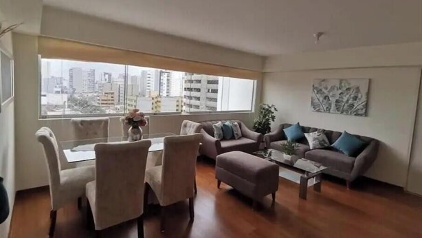 A spacious living room with a dining area, large windows with a city view, and a modern interior design.