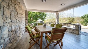 A covered outdoor dining area with stone walls, a table, chairs, and a view of trees and hills.