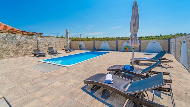 A rooftop pool area with lounge chairs and umbrellas, surrounded by stone walls and a clear blue sky.