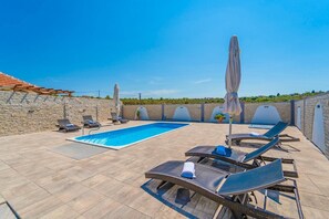 Zona de piscina en la azotea con sillones de descanso y sombrillas, rodeada de muros de piedra y un cielo azul despejado.