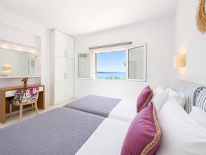 A bedroom with a bed, a desk, a chair, a wardrobe, and a window with a view of the sea.