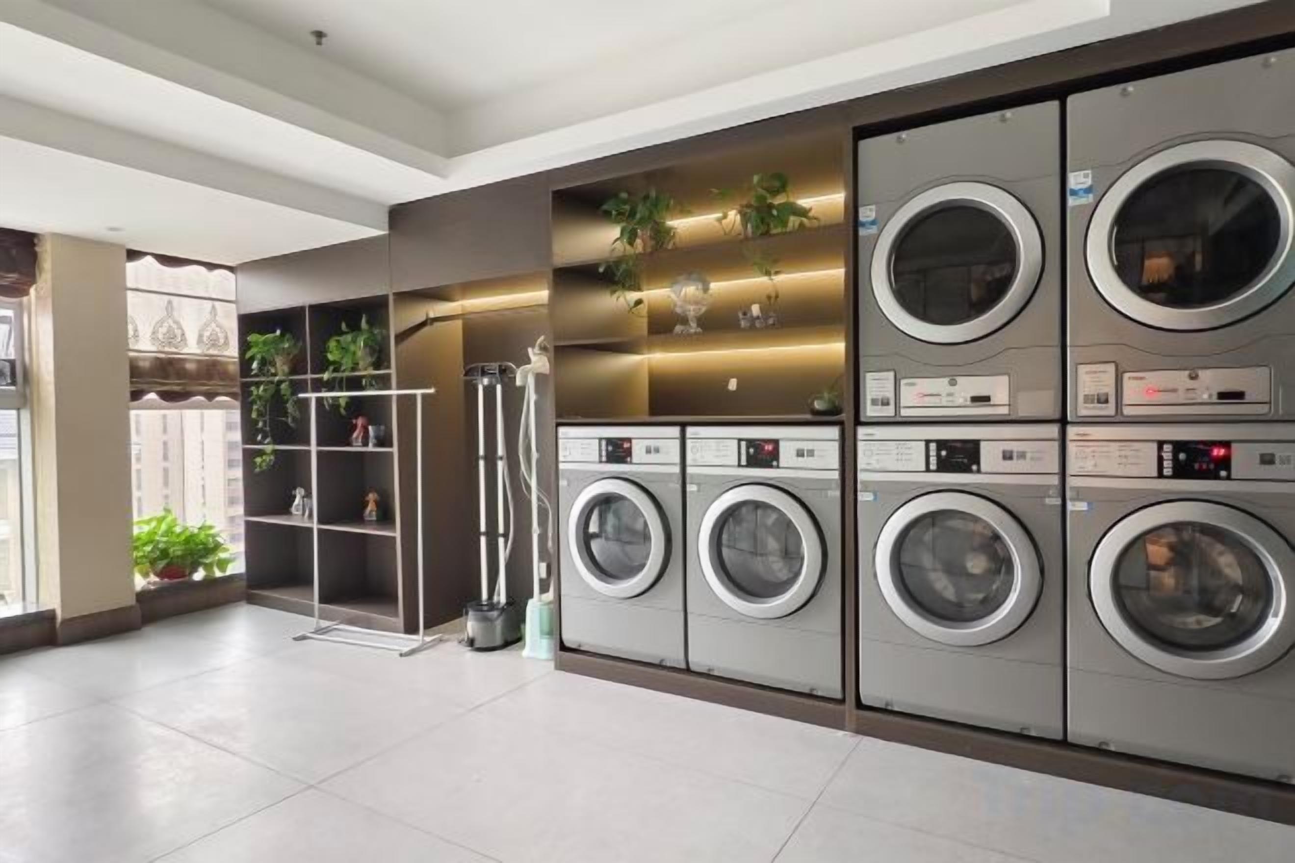 A laundry facility with multiple washing machines and dryers, a metal drying rack, and shelves with plants.