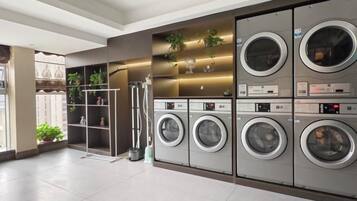 A laundry facility with multiple washing machines and dryers, a metal drying rack, and shelves with plants.