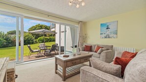 A spacious living area with a sliding glass door leading to a patio, a wooden coffee table, a grey sofa, and a painting of a lighthouse on the wall. - Harbour Reach - Close to Beach, Parking (Saundersfoot)