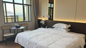 A hotel room with a large bed, a small table with chairs, a nightstand, and a view of the sea through a large window.