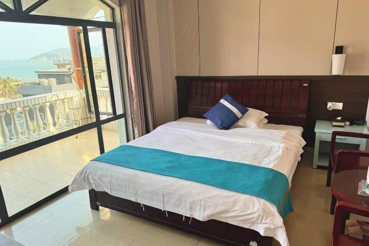 Ein Hotelzimmer mit großem Fenster, einem Bett mit weißer Bettwäsche, einem blauen Kopfkissen, einem Holz-Kopfteil, einem Nachttisch, einem Schreibtisch und einem Stuhl.