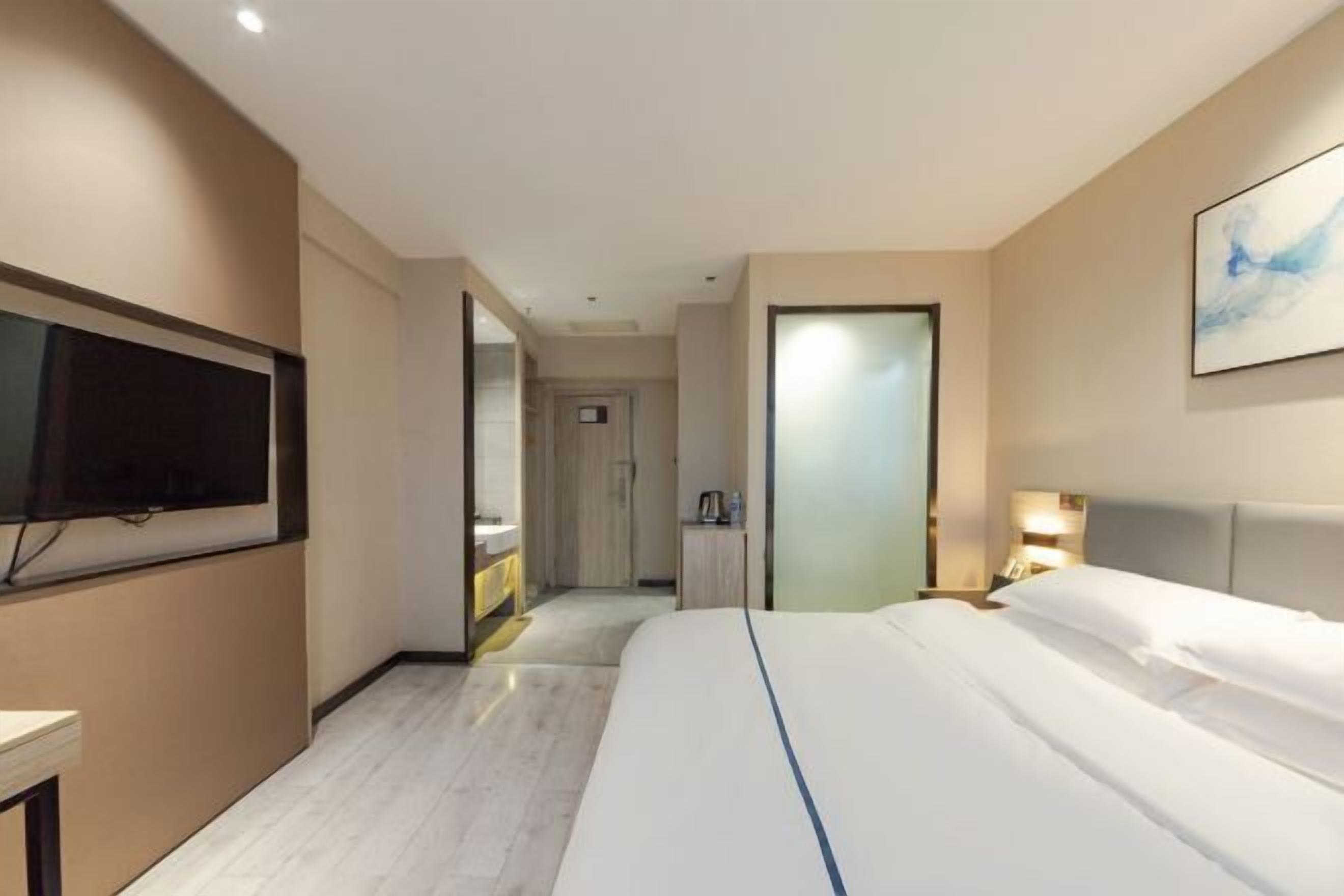 A hotel room with a large bed, a flat-screen TV mounted on the wall, a bedside table with a lamp, and a door leading to a bathroom.