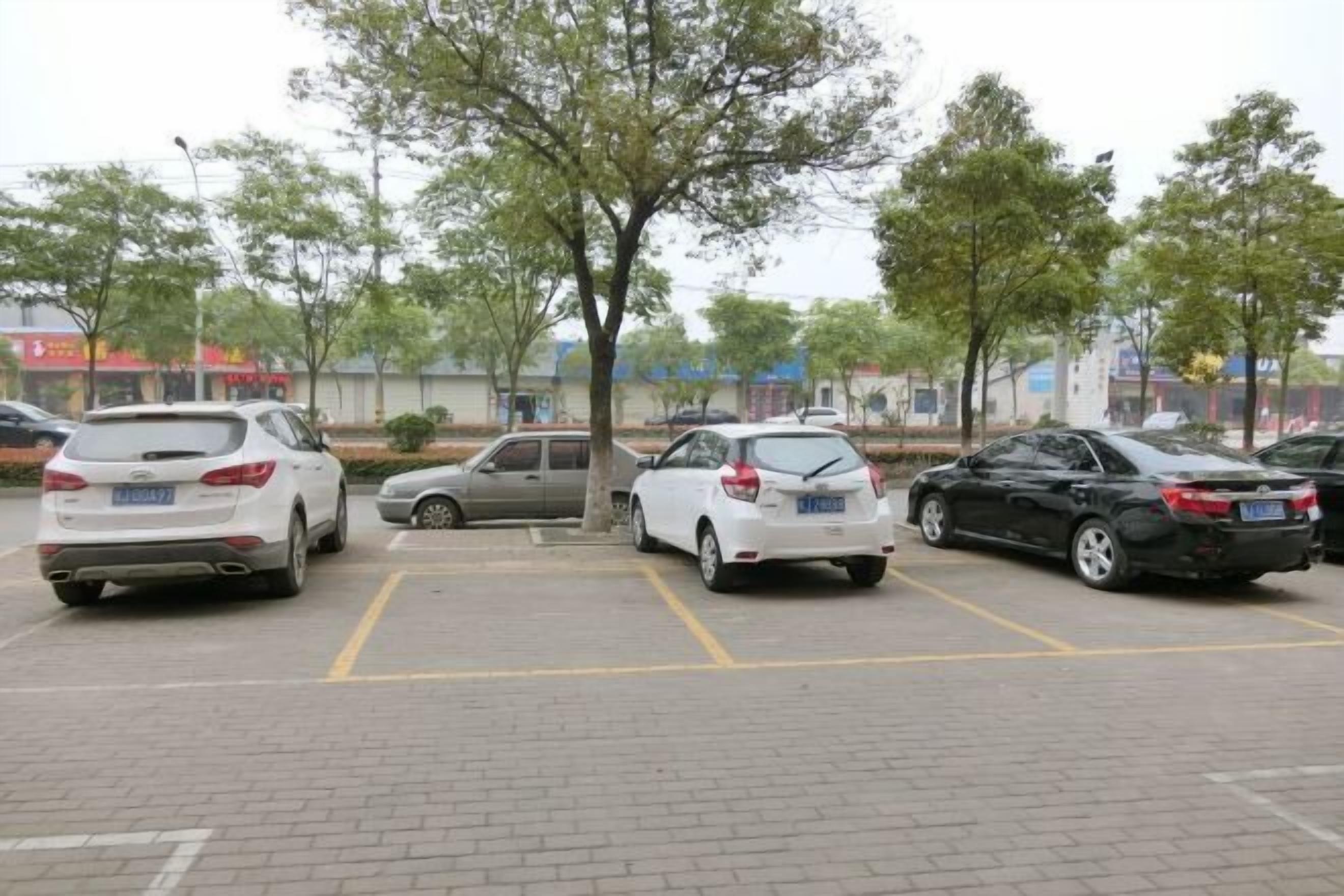 A parking lot with several cars parked, trees, and buildings in the background.