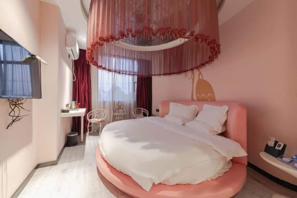 Une chambre avec un lit rose, du linge de lit blanc, un bureau avec une chaise et un téléviseur fixé au mur.