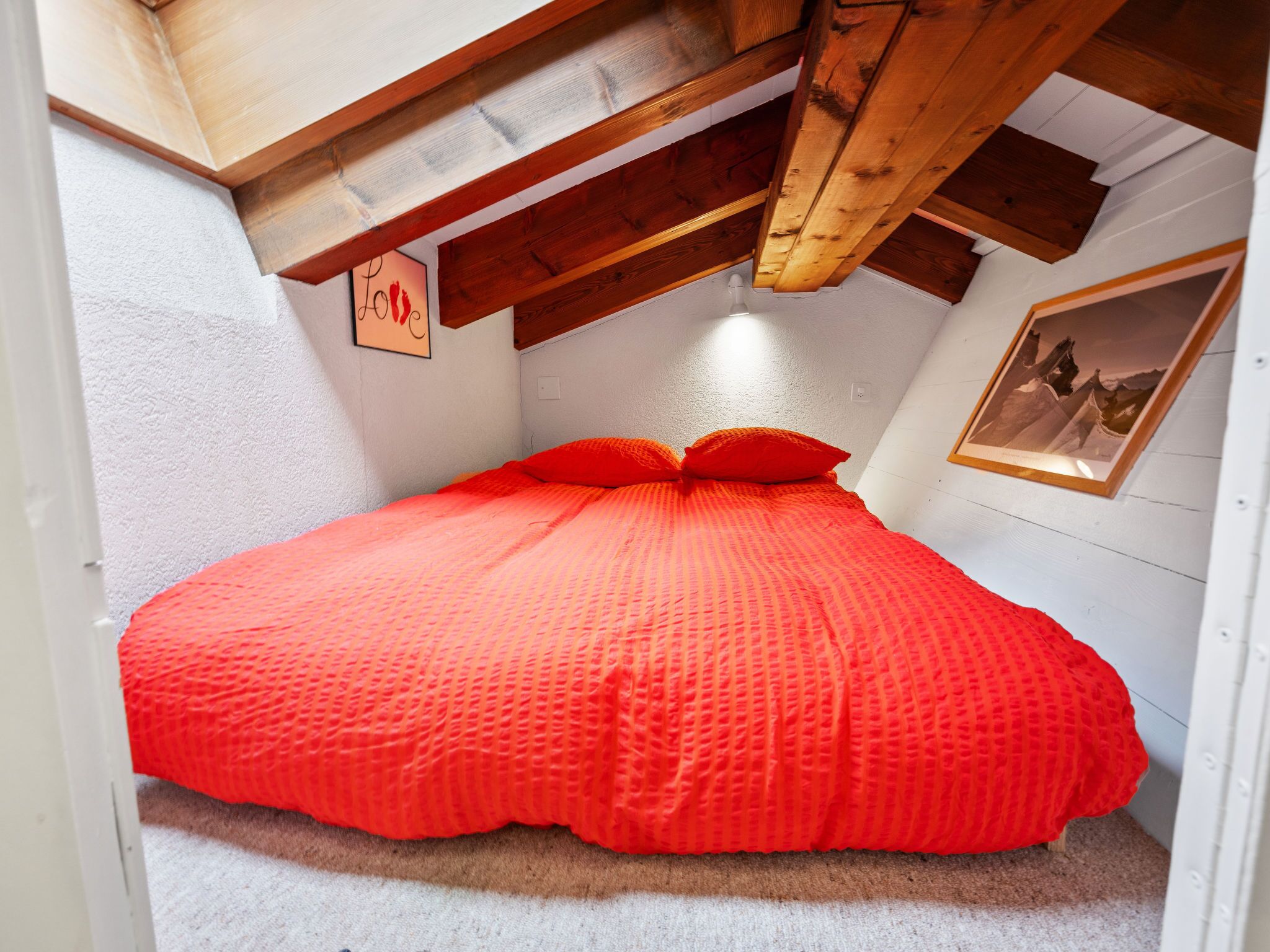 Un dormitorio pequeño y acogedor con techo inclinado y vigas de madera, que presenta una cama individual con una colcha y almohadas de color naranja brillante, un cuadro enmarcado en la pared y una pared blanca con textura.