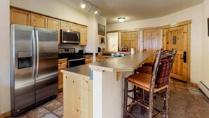 A kitchen with a stainless steel refrigerator, microwave, and oven. There is a wooden bar counter with two bar stools. The kitchen has wooden cabinets and a tiled floor. - Tenderfoot Lodge 2 Bedroom at the Mountain House Base (Keystone)