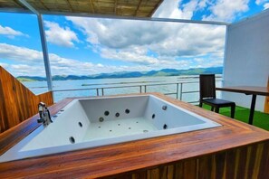 Une terrasse en bois avec une grande baignoire et une table, offrant une vue sur une étendue d’eau et des montagnes lointaines.