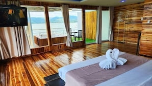 A hotel room with a wooden floor, a bed with a white comforter and a swan-shaped towel, a TV on the wall, and a balcony with a view of the water. - Banrai WanWan Resort (Si Sawat)