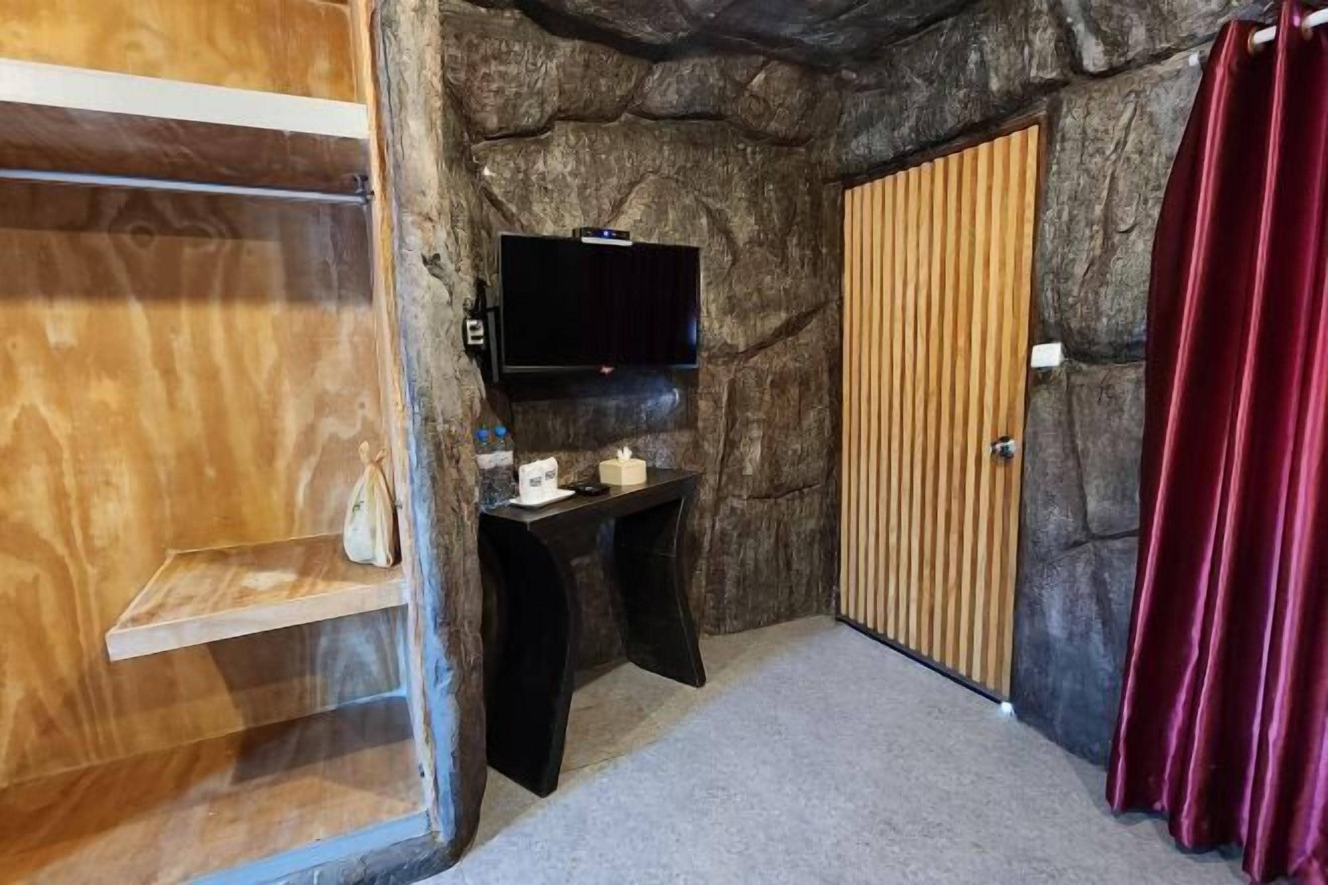 Una habitación con pared de piedra, puerta de madera, televisor negro sobre un soporte, una mesita con una caja de pañuelos y una cortina roja.