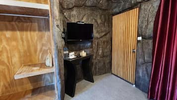 Una habitación con pared de piedra, puerta de madera, televisor negro sobre un soporte, una mesita con una caja de pañuelos y una cortina roja.