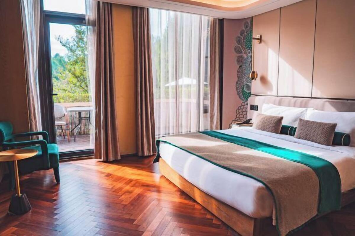 Ein Hotelzimmer mit einem großen Bett, einer Sitzecke mit einem Sessel, Blick durch eine Glastür und einer dekorativen Wandverkleidung.