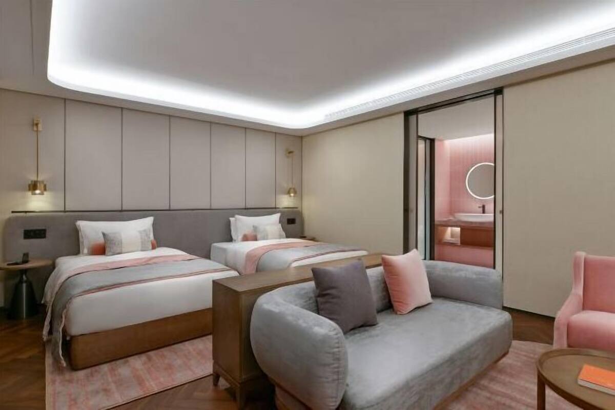 Ein modernes Hotelzimmer mit zwei Betten, einem Schlafsofa, einem kleinen Tisch und einem durch eine Schiebetür einsehbarer Badezimmer.