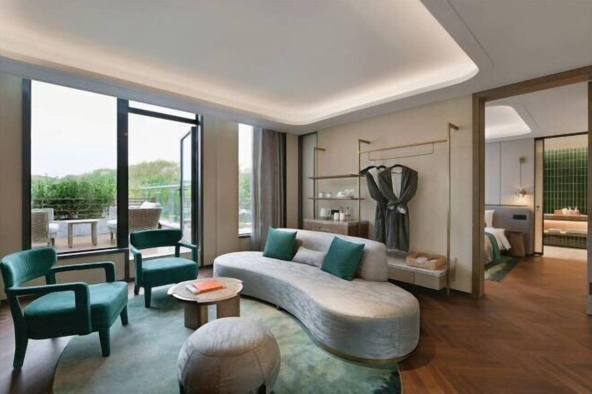 Ein modernes Hotelzimmer mit einer gebogenen Couch, grünen Sesseln, einem kleinen runden Tisch und Blick ins Grüne durch große Fenster.