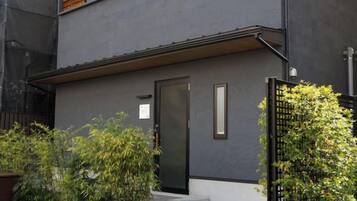 Un edificio moderno de dos pisos con fachada oscura, una puerta negra y un ventanal pequeño. Hay escalones que conducen a la entrada, y un portón negro con plantas a la derecha.