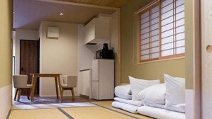 Une pièce de style japonais traditionnel, avec un sol recouvert de tatamis, une petite table basse entourée de chaises, un lit avec une literie blanche et une fenêtre coulissante de type shoji.