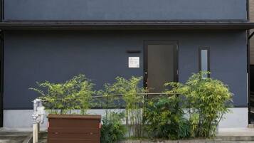 Un edificio de fachada oscura, una puerta y una pequeña ventana. Hay un contenedor marrón y algo de vegetación al frente.
