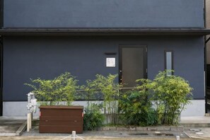 A building with a dark facade, a door, and a small window. There is a brown storage unit and some greenery in front. - Guesthouse ZenNichi (Kyoto)