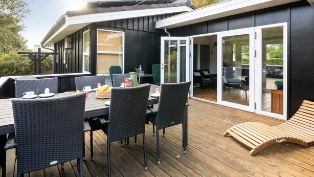 Un comedor moderno al aire libre con terraza de madera, mobiliario de mimbre negro y puerta de cristal que da a un espacio interior.