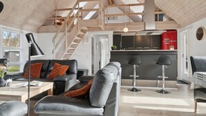 Un espacio habitable moderno estilo loft con techo alto, vigas de madera a la vista, una escalera, zona de cocina y zona de estar con sofás de cuero y una mesa de centro.