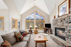 A high-ceilinged living room with a stone fireplace, large windows, a sofa, and a television. - 4 Bedroom Condo at Gateway Lodge Near Mountain House Base (Keystone)