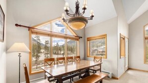 A dining area with a large wooden table, six chairs, a bench, a chandelier, and large windows with mountain views. - 4 Bedroom Condo at Gateway Lodge Near Mountain House Base (Keystone)