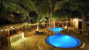 Un área de piscina exterior de noche con dos piscinas, sillones reclinables y luces colgantes.