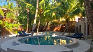 Una piscina circular con sillas de descanso, rodeada de palmeras y una estructura con techo de paja.
