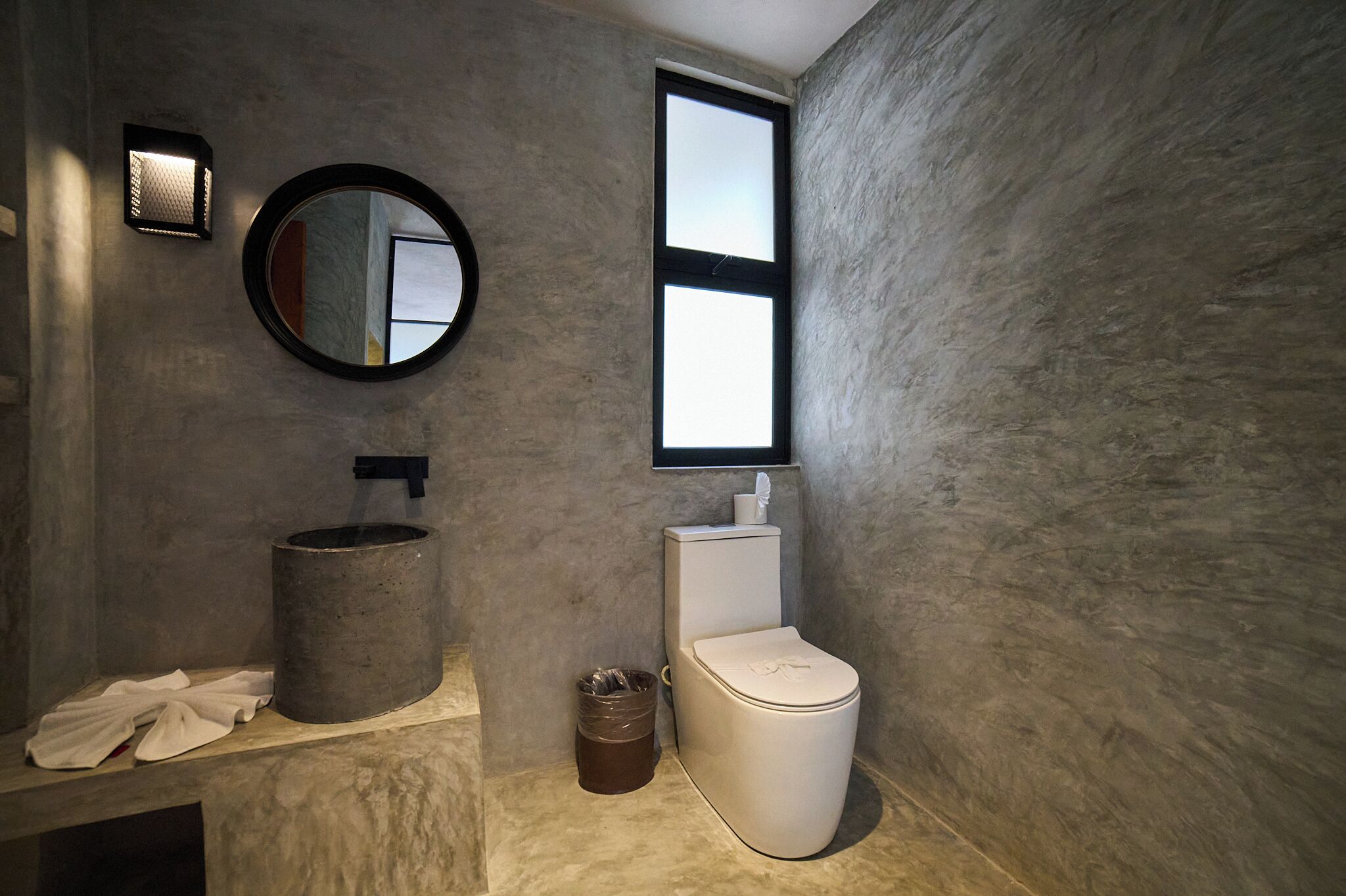 A modern bathroom with a round mirror, a cylindrical sink, a toilet, and a window.