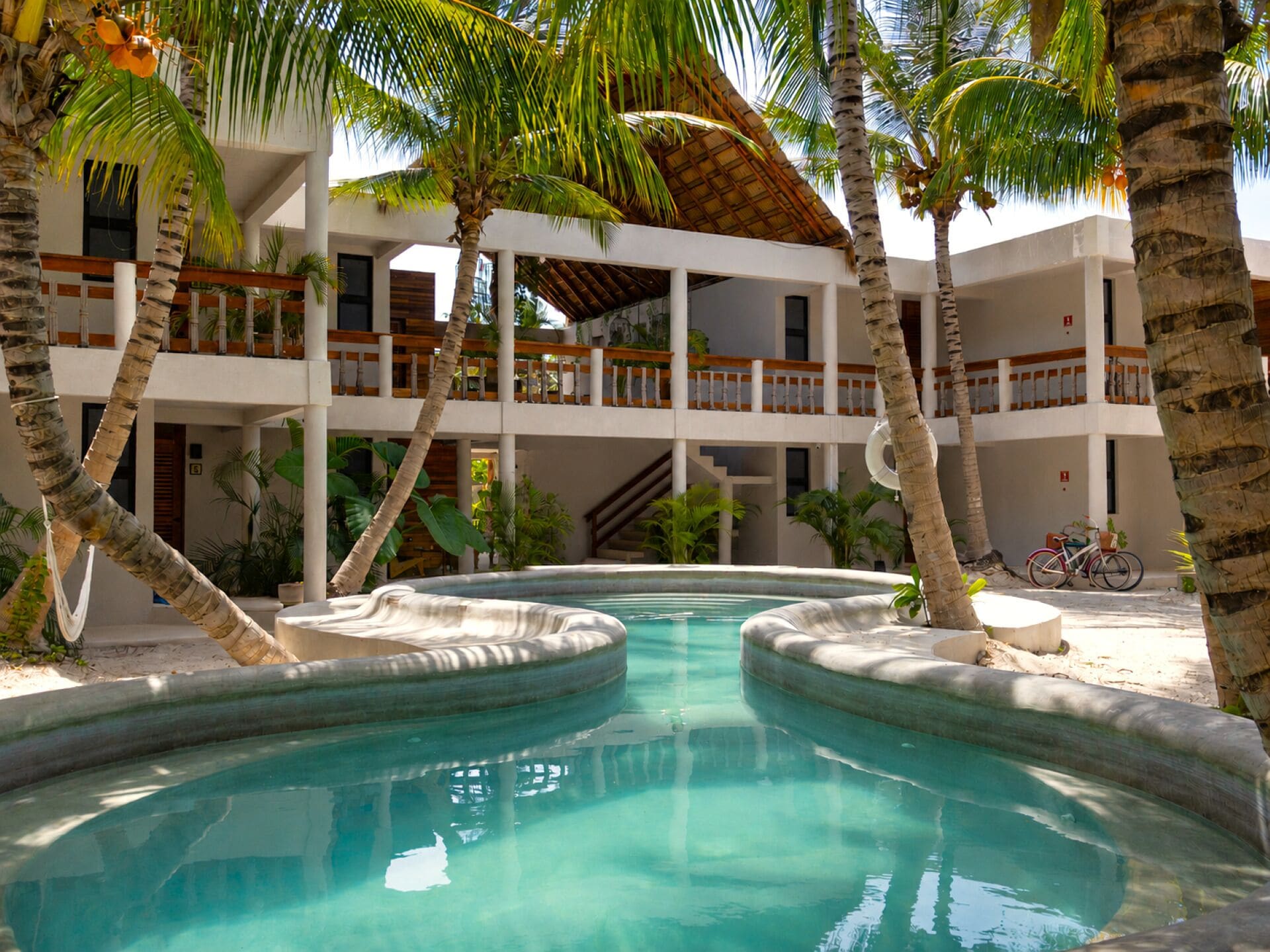 Una piscina con diseño circular, rodeada de palmeras y un edificio de varios niveles con balcones.