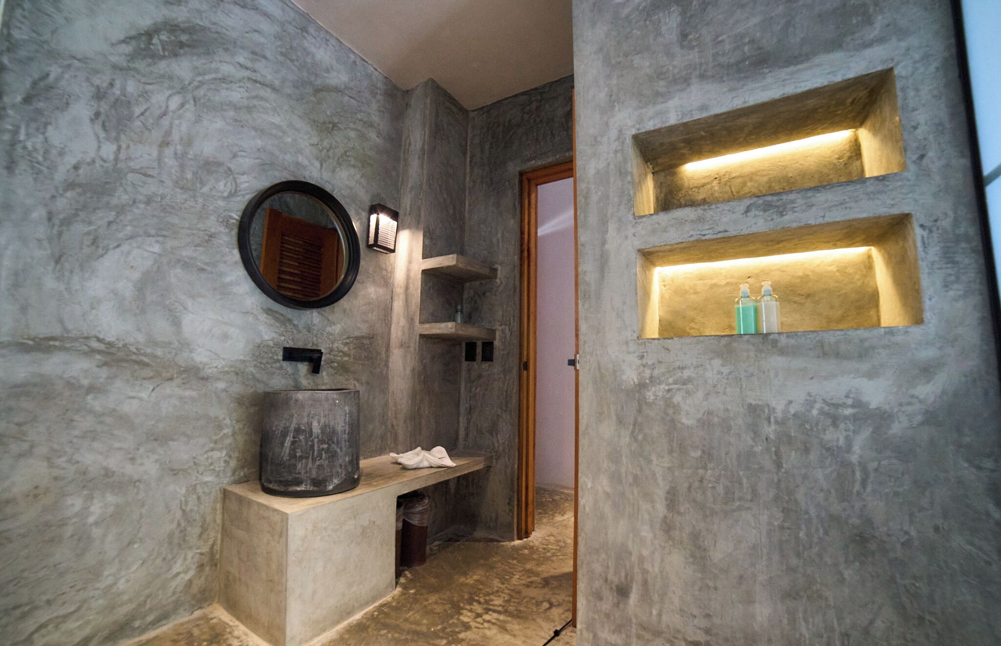 A bathroom with a marble wall, a round mirror, a sink with a basin, and two illuminated shelves.