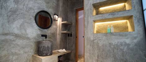 A bathroom with a marble wall, a round mirror, a sink with a basin, and two illuminated shelves.