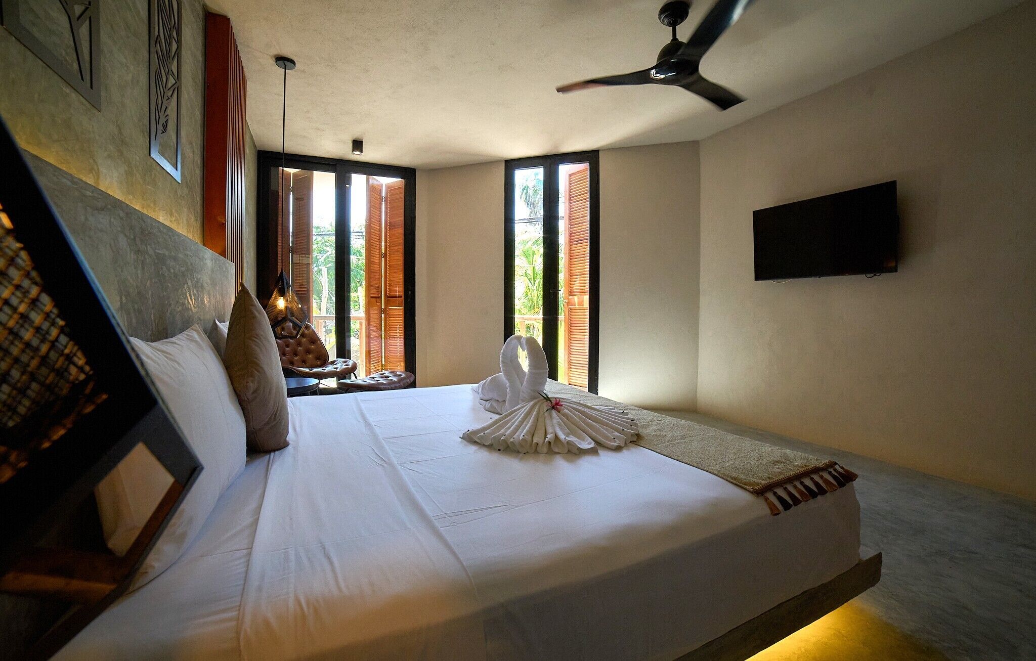 A hotel room with a bed, a ceiling fan, a television, and a view of the outdoors through the windows.
