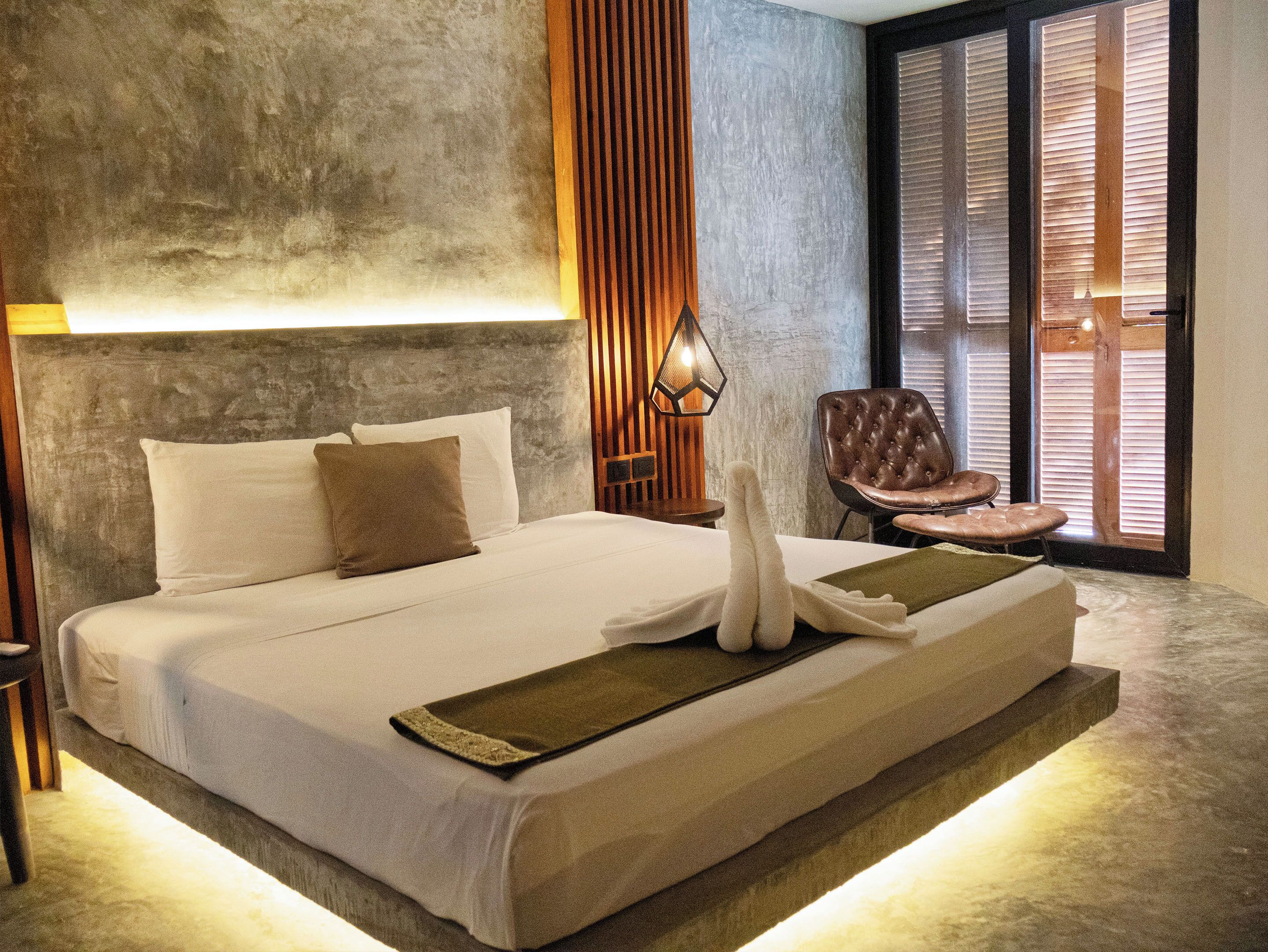 A bed with white linens, a wooden headboard, a wall-mounted lamp, and a chair with a cushioned seat.