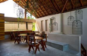 Un interior con zona de comedor, muebles de madera, techo de paja y adornos de jaulas de pájaros colgadas en las paredes.
