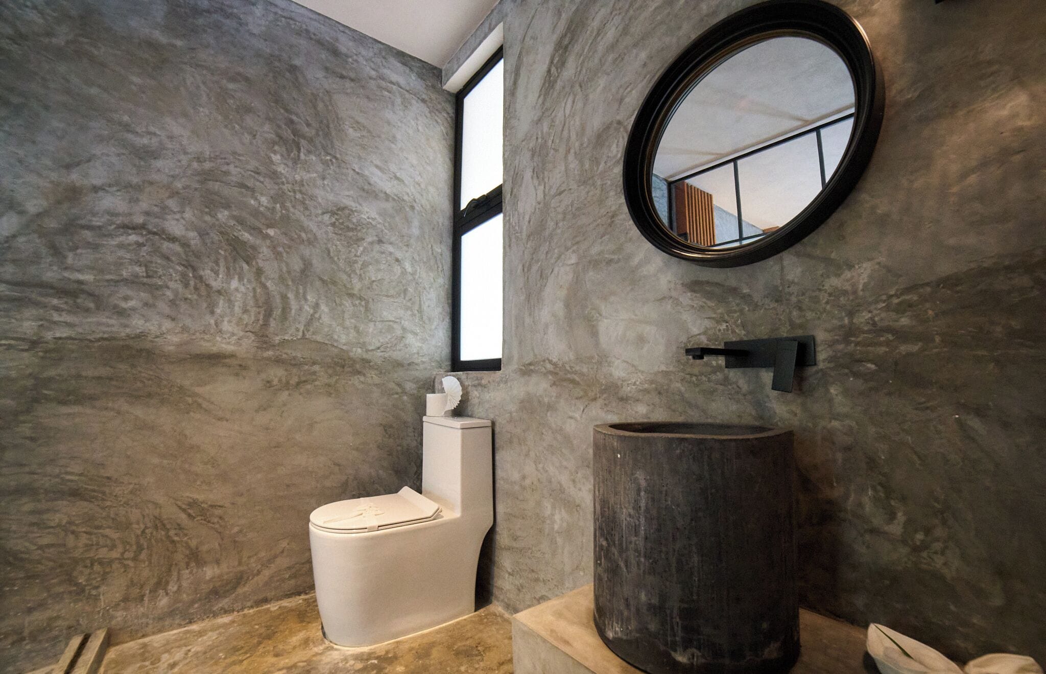 Une salle de bain avec un mur en marbre, un miroir rond, des toilettes et un grand lavabo en bois.