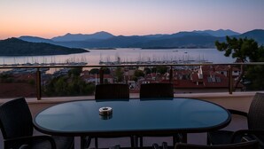 A round table with four chairs, a small table lamp, and a view of a marina with boats and a mountainous landscape in the background.