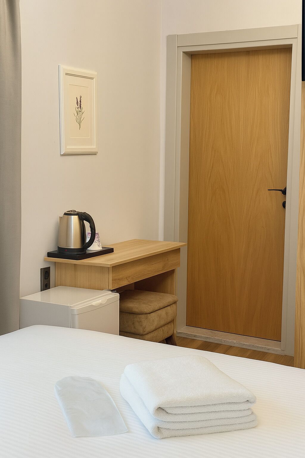 A hotel room with a bed, a wooden desk, a kettle, and a framed picture on the wall.