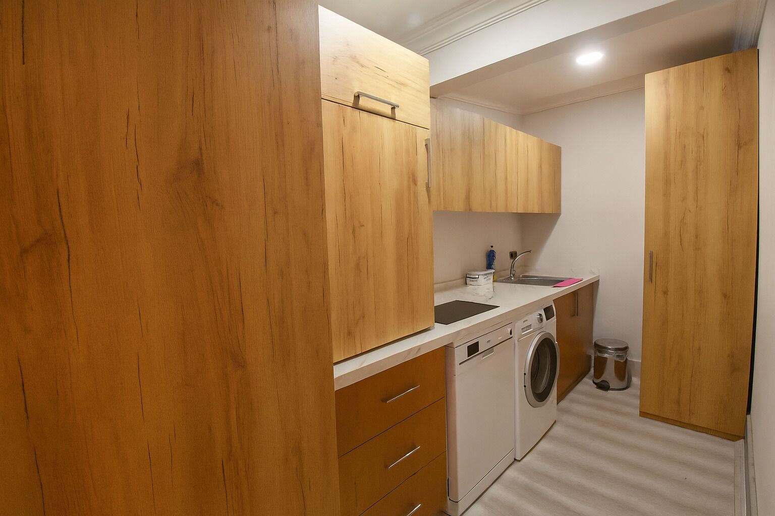 A compact kitchen with wooden cabinets, a washing machine, and a small trash bin.